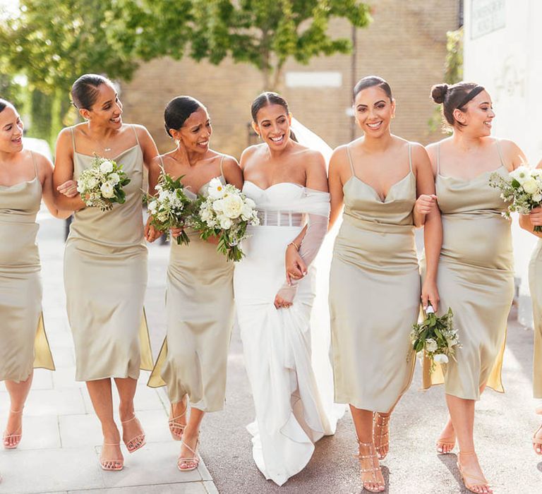 Bridesmaids with nude heels and sage green cowl neck satin dresses walk the streets together with bride 