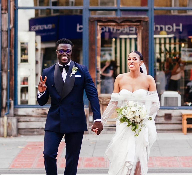 Groom in blue and black tuxedo throws up a peace sign as he crosses the road with bride in pink wedding shoes 