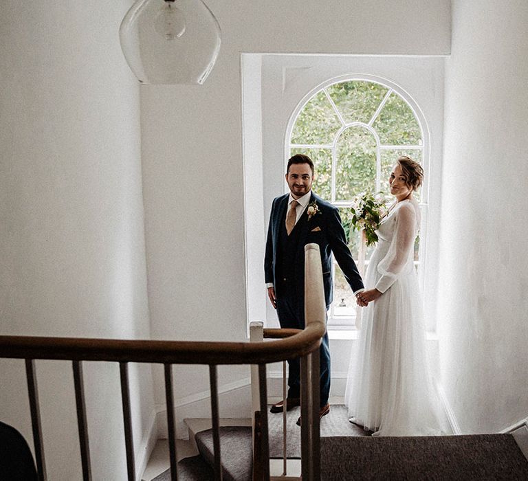 Bride in sheer long sleeve wedding dress with tulle skirt standing with groom in blue suit with gold patterned tie