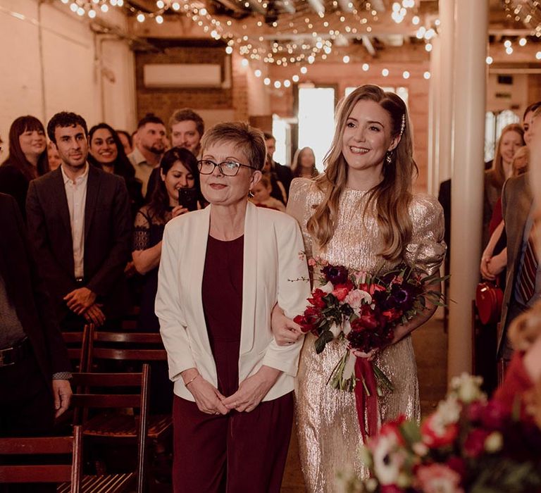 Mother of the bride in burgundy dress and white blazer walks the bride in a gold wedding dress down the aisle 