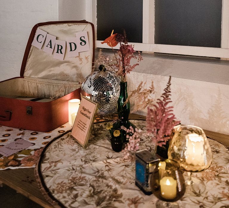 Open suitcase for wedding cards and guestbook with vases of flowers and candles with disco ball and sign in gold photo frame