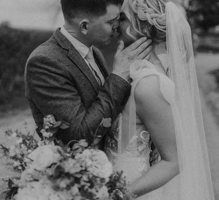 Bride with blonde hair in braided ponytail with diamanté hair clip kissing groom holding wedding bouquet