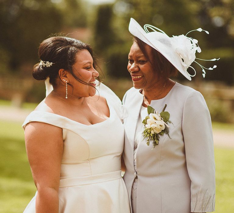 Mother of the bride in grey outfit and hat smiles with the bride in bardot style wedding dress 