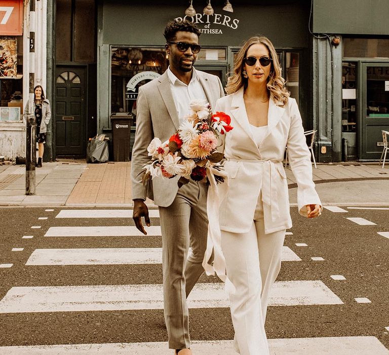 Bride in a trouser suit and sunglasses crossing the road with her groom at their Portsmouth Guildhall wedding 