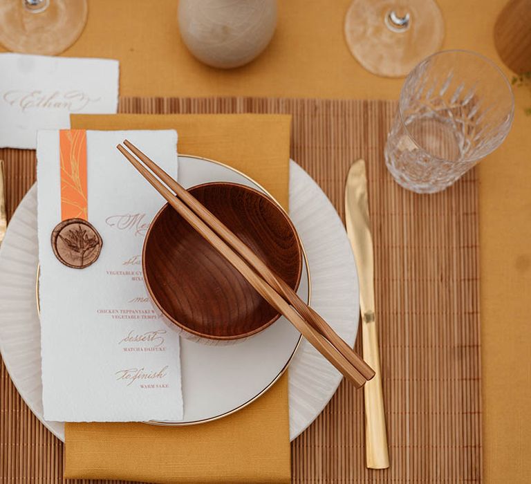 Place setting with yellow table rubber, bamboo place mat, gold cutlery, brown rice bowls, wooden chopsticks and menus card for Japanese wedding 