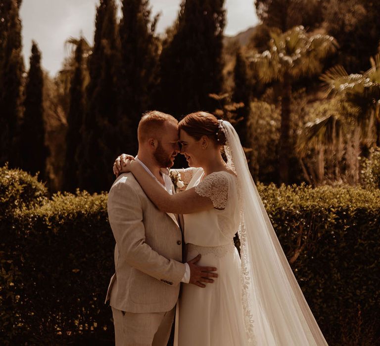 Bride wears gown complete with lace sleeve detailing and floor length veil with lace trim 