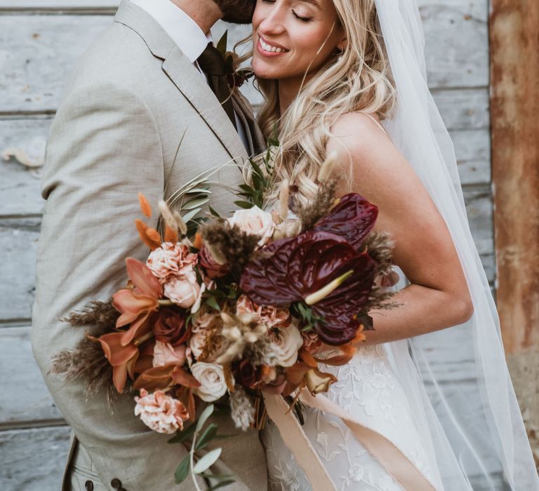 Groom kisses his bride as she holds bridal bouquet filled with orange orchids, bunny ears, and deep red anthurium 