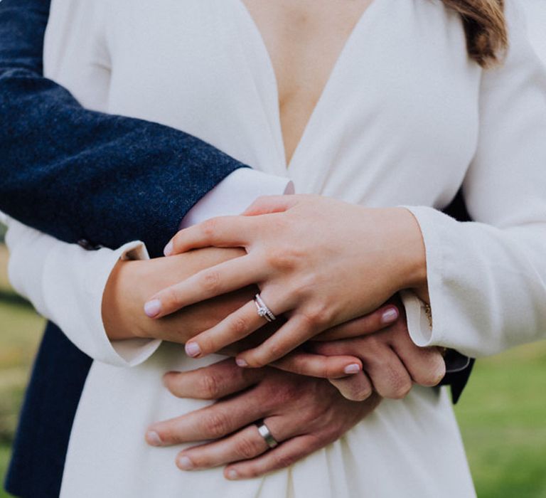 Bride with pale pink nails and an round engagement ring and wedding band with the groom showing his plain wedding ring 