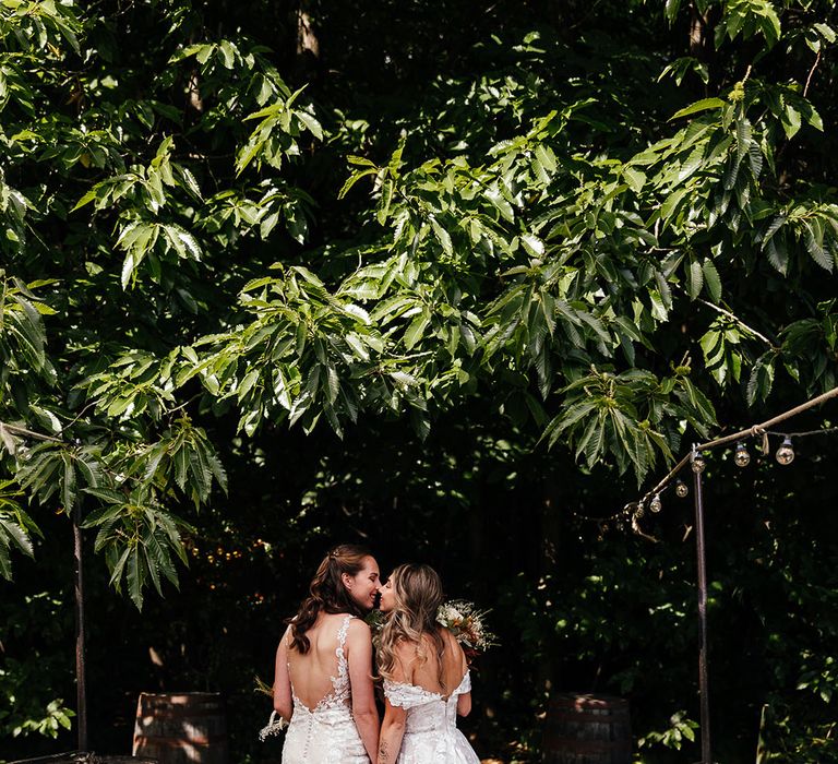 Bride in illusion back button wedding dress and bride in off the shoulder wedding dress lean in for a kiss together 