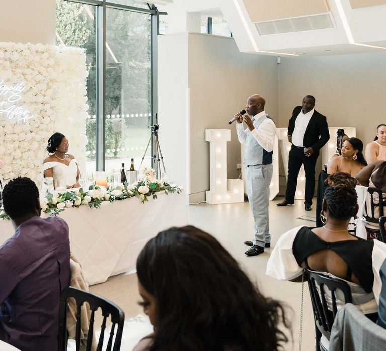 Bride & groom sit in front of white rose flower wall with neon sign as guest gives speech during reception 