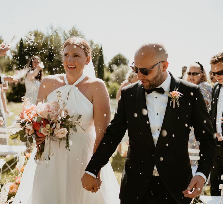 Bride wearing Halfpenny London wedding dress walks through confetti with her groom in black tie and sunglasses 