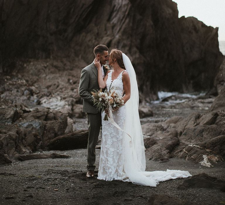 Bride holds neutral toned bridal bouquet tied with ribbon as she leans in to kiss her groom at Tunnels Beach