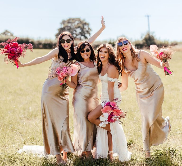 Bride wearing off-the-shoulder lace wedding dress stands with her bridesmaids who wear satin champagne bridesmaid dresses and all hold bright pink floral bouquets