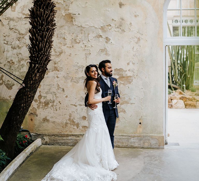 Bride & groom stand beside rustic wall at the Great Conservatory in Syon Park