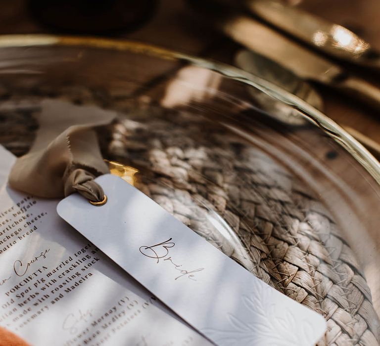 Gold rimmed clear charger plates with wicker placemats and minimal style place names and menu