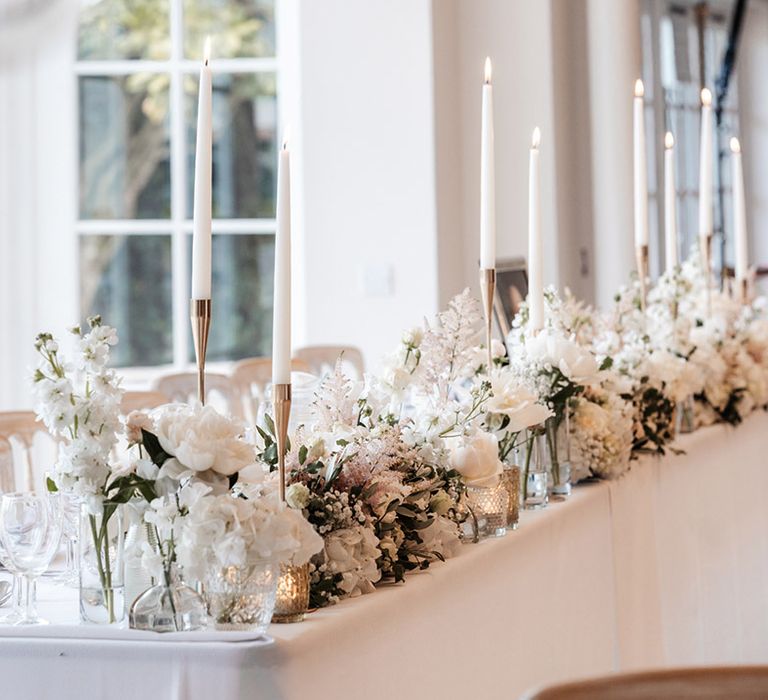 White floral centrepieces line banquet table with white tablecloth and white candles in gold holders