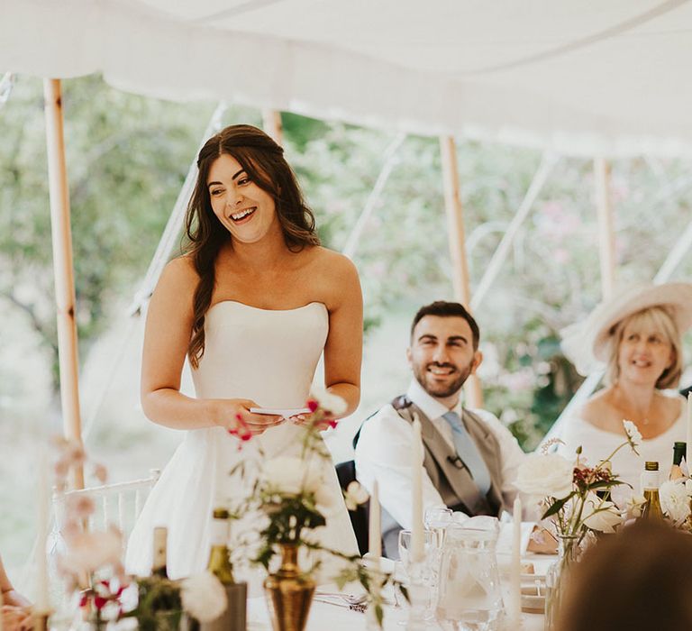 Bride in a fitted strapless wedding dress stands up as she delivers a bridal speech