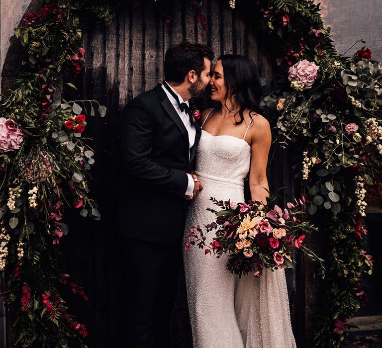 The bride in a sparkly wedding dress smiles with the groom in black tie under the pink, red and purple wedding flower arch 