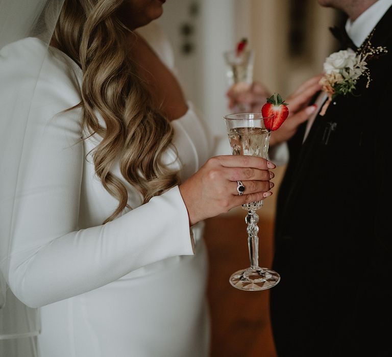 Bride in long sleeve satin wedding dress with silver wedding ring and black stone and groom in black suit with floral boutonniere holding Champagne glasses with strawberry slices 