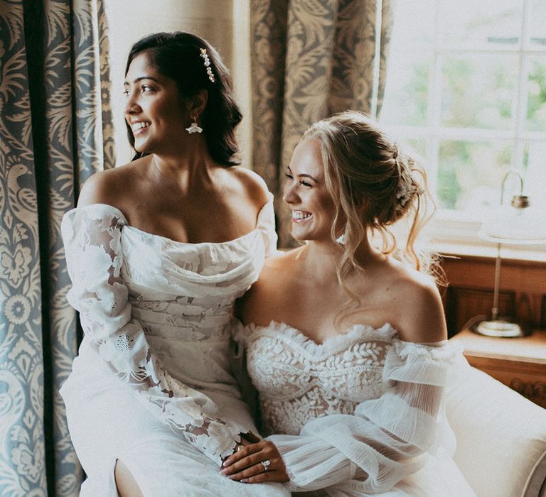 Bride in off the shoulder long sleeve wedding dress sitting on bride's lap wearing strapless lace wedding dress on cream coloured chair