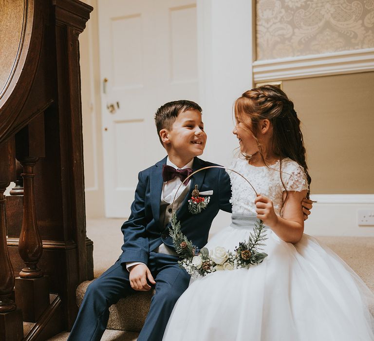 Page boy in a blue suit with burgundy bow tie sitting with the flower girl in a white dress with white lace detailing 