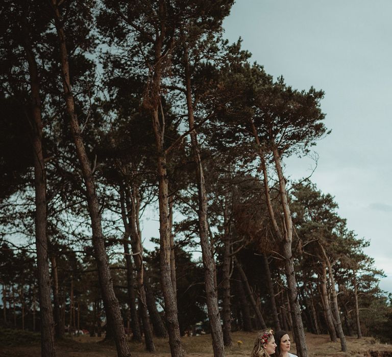 Brides stand together with flower hair accessories looking out at the beach 