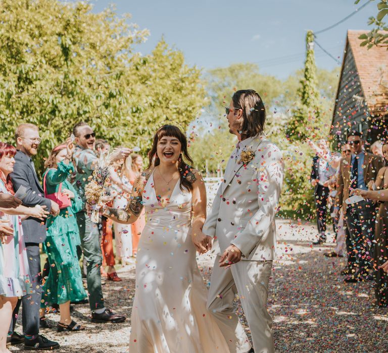 Groom in neutral coloured double-breasted ASOS suit and black high-top converse with dried flower and foliage boutonniere holding hands with bride in silk-look v-neck sleeveless ASOS wedding dress doing confetti walk 