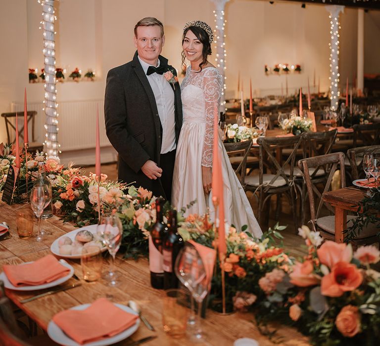 Bride in taffeta material princess wedding dress with long lace sleeves  and groom in black tux standing in reception room with silver and gold streamer decorations and fairy lights 