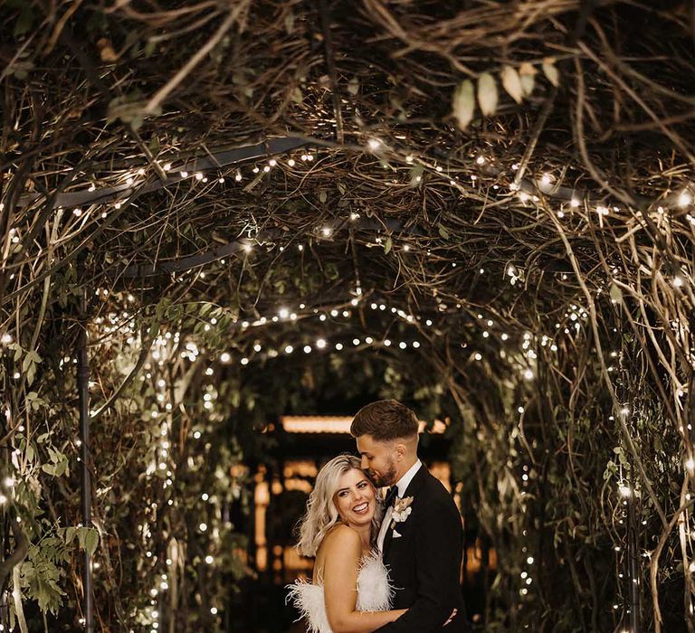 Bride in strapless white satin look feather jumpsuit and groom in classic Ralph Lauren black tuxedo with black bowtie and dried flower boutonniere embracing in tunnel of foliage and fairy lights at The Old Kent Barn wedding venue