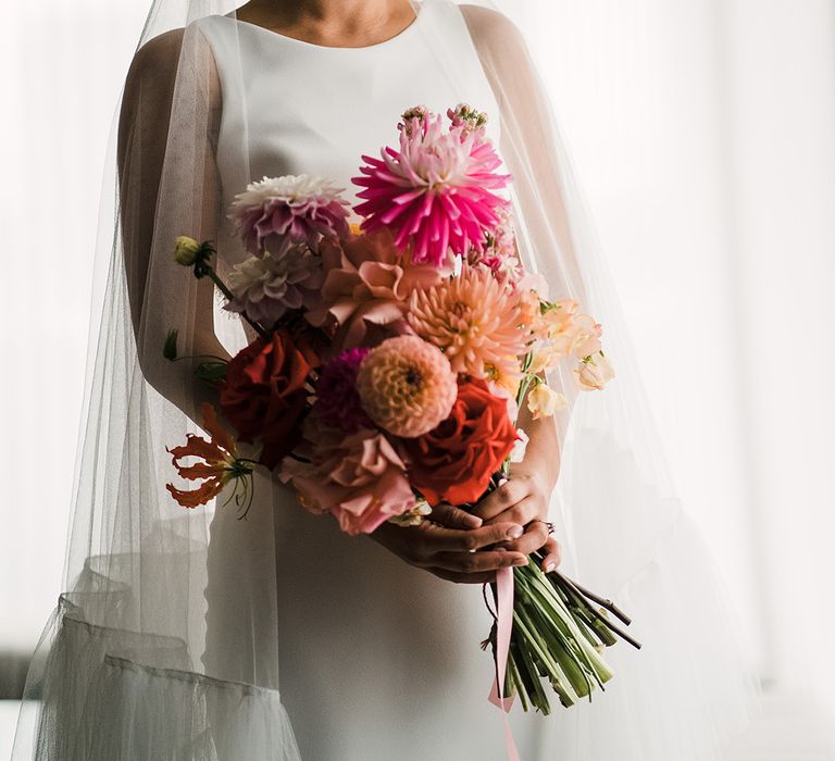 East Asian bride holds colourful floral bouquet and wears fitted wedding dress with bateau neckline and embellished bridal crown with ruffle edge drop veil