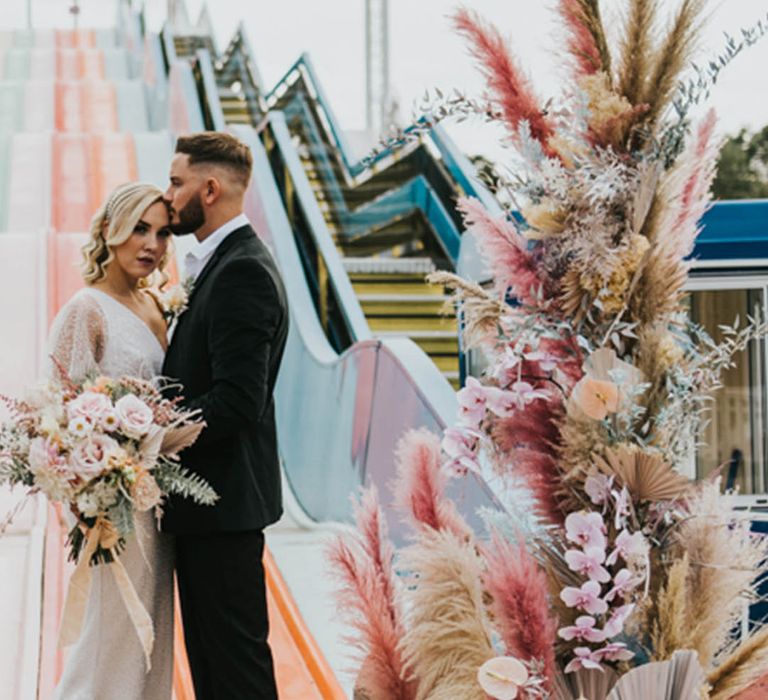 Fun theme park theme wedding with colourful pampas grass boho wedding flower column arrangement with the bride and groom next to colourful slides