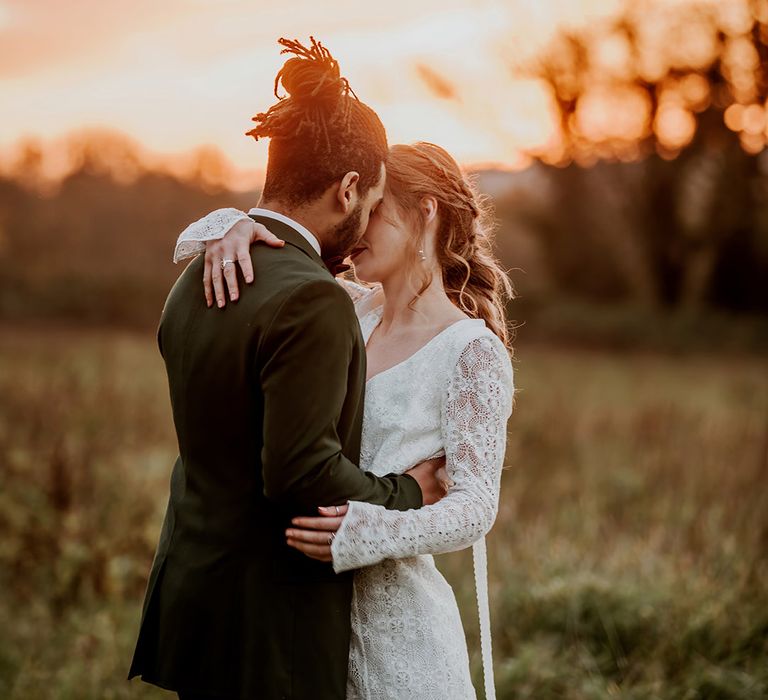 Groom in classic black tux embracing bride in boho long sleeve lace wedding dress at golden hour photo shoot 