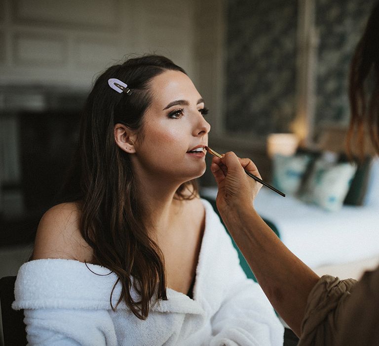 Bride sits in a white robs getting ready for the wedding day as she has her makeup done 