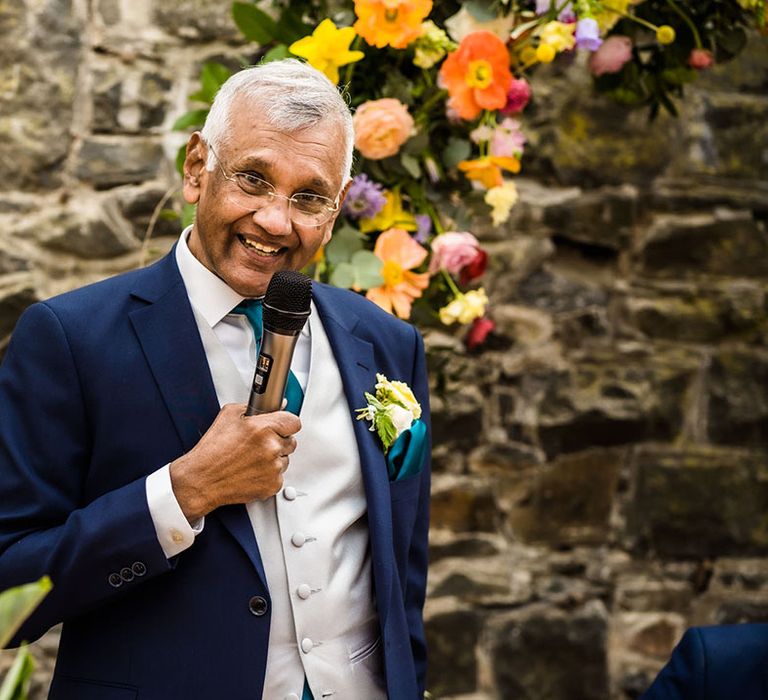 Brides father gives speech in front of colourful floral arch 