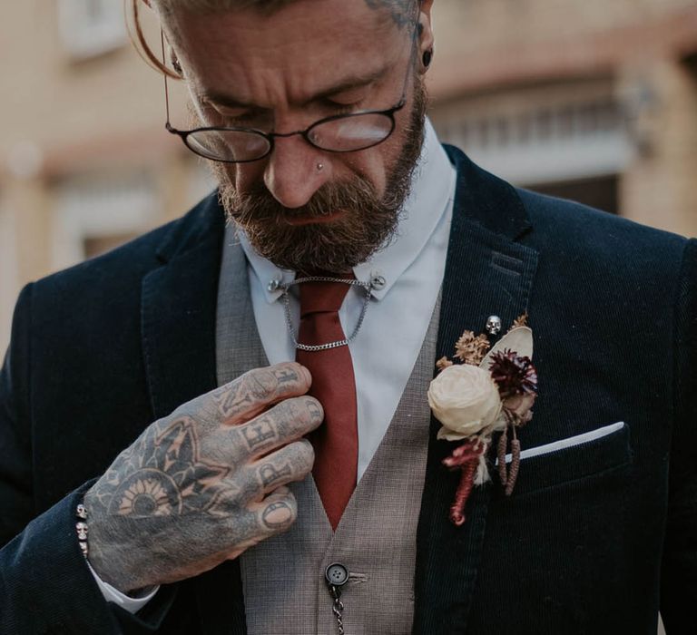 Tattooed groom in dark suit jacket with a grey waistcoat and orange tie with silver chain and jewellery with white rose buttonhole and skull pin 