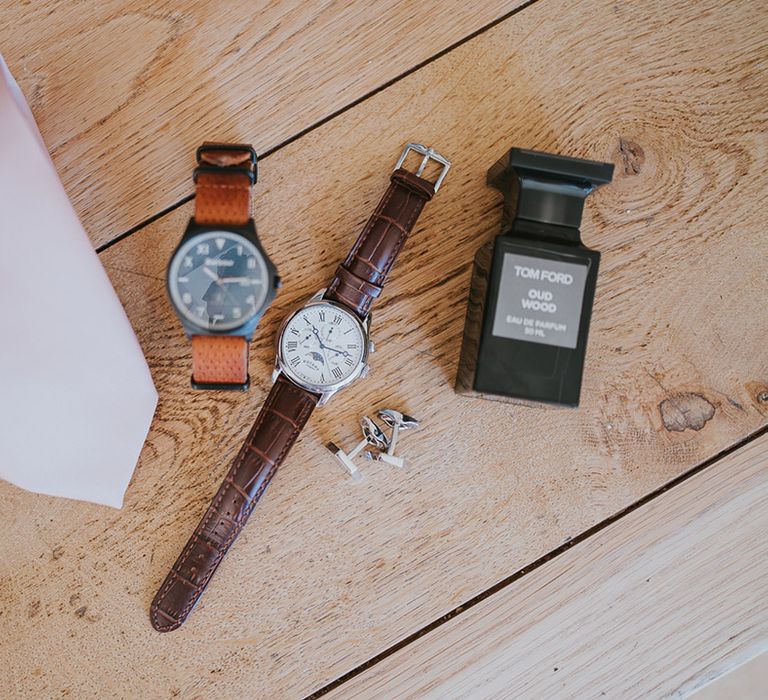 Grooms accessories with Tom Ford cologne, a pink tie, silver cufflinks, a brown leather watch, and a woven watch 