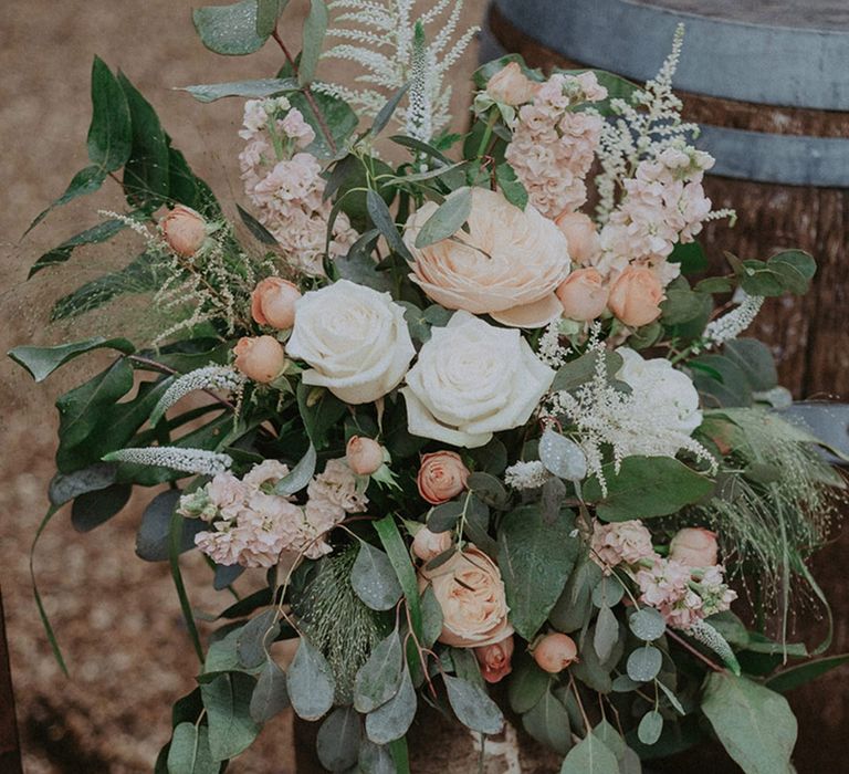 Pale pink and peach roses, peonies, stock and more flowers sit with foliage for the wedding flower decor and arrangements at the rustic luxe Stanlake Park wedding