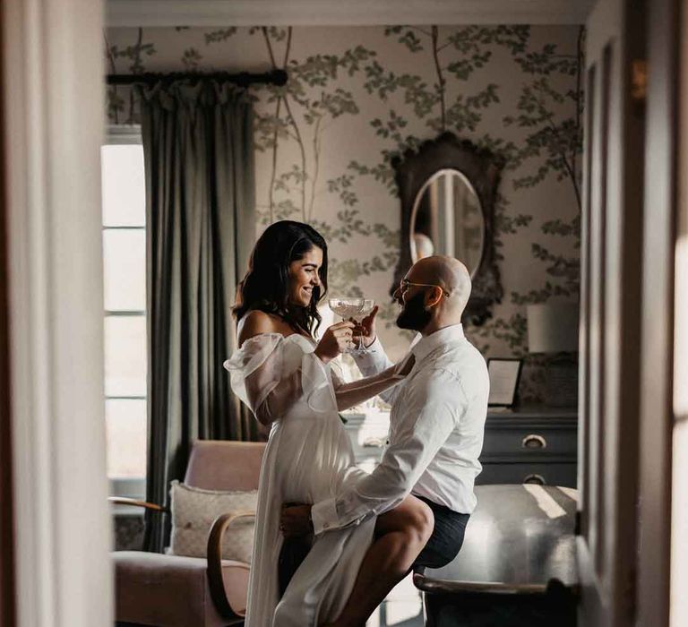 Bride in sheer puff sleeve wedding dress with front slit embracing groom in white shirt and black suit trousers, sharing a drink together 