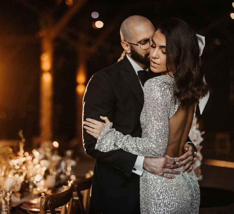 Bride in long sleeve silver sparkly wedding dress with open back detail embracing groom in classic black tuxedo 