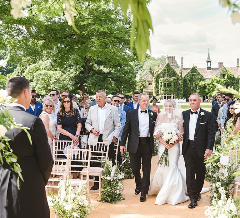 Two men in black tie walk the bride in a fitted wedding dress down the aisle for the outdoor wedding ceremony 