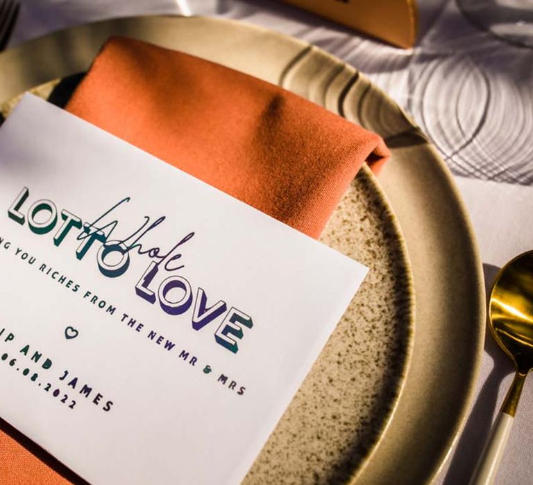 White and metallic holographic wedding place setting welcome card on orange napkin and speckled plates with gold cutlery 