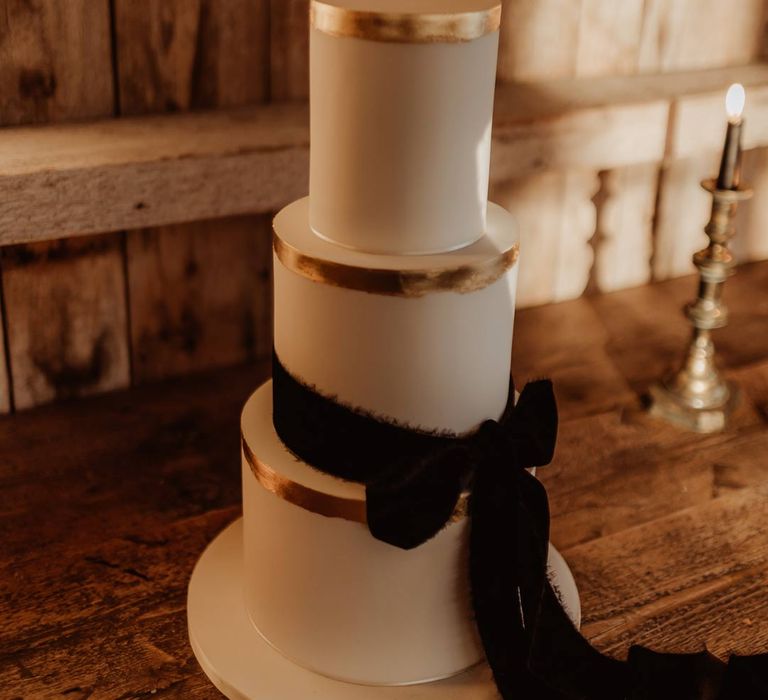 Three tiered buttercream wedding cake with gold rim and large black velvet bow tied around the front on wooden table at rustic wedding 