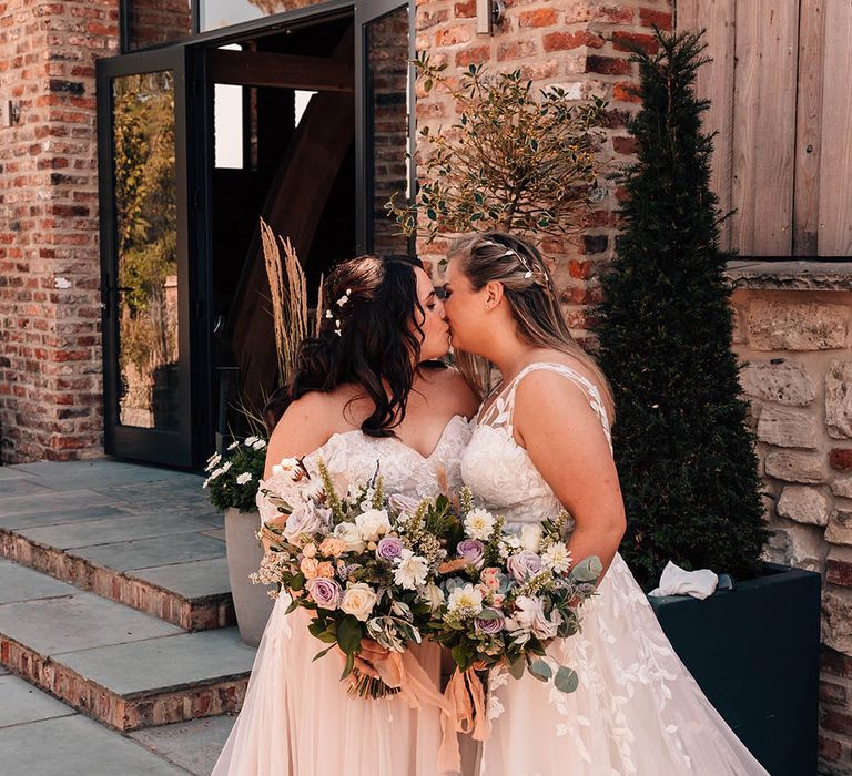 Brides wearing pretty floral lace wedding dresses share a kiss holding matching purple ad white bouquets tied with pink ribbon 