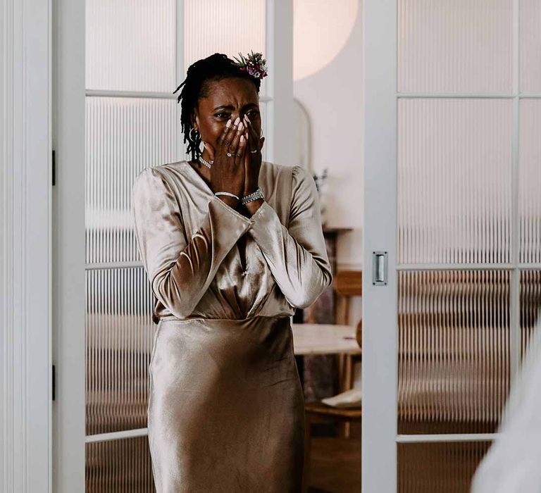 Mother of the bride wearing long sleeve golden silk wrap around dress and black fascinator doing bridal reveal 