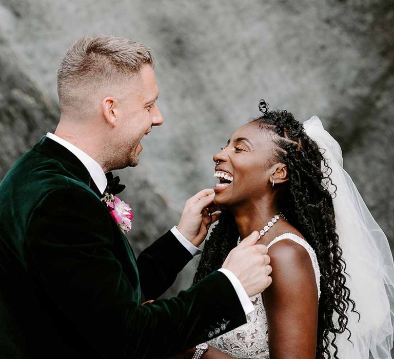 Groom in velvet grooms blazer, colourful tropical boutonniere and black suit trousers laughing with bride in v neck sleeveless lace Pronovias wedding dress, cathedral length veil and silver sparkly bridal jewellery on Tunnels Beaches 