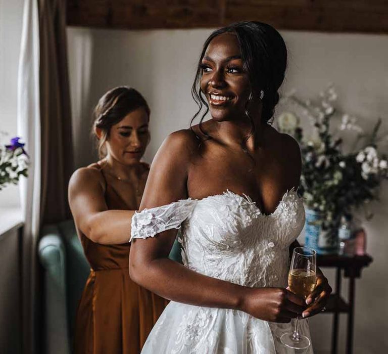 Bridesmaid in burnt orange bridesmaid dress helping bride with bridal preparations in her sweetheart neckline lace wedding dress with detachable off the shoulder sleeves 