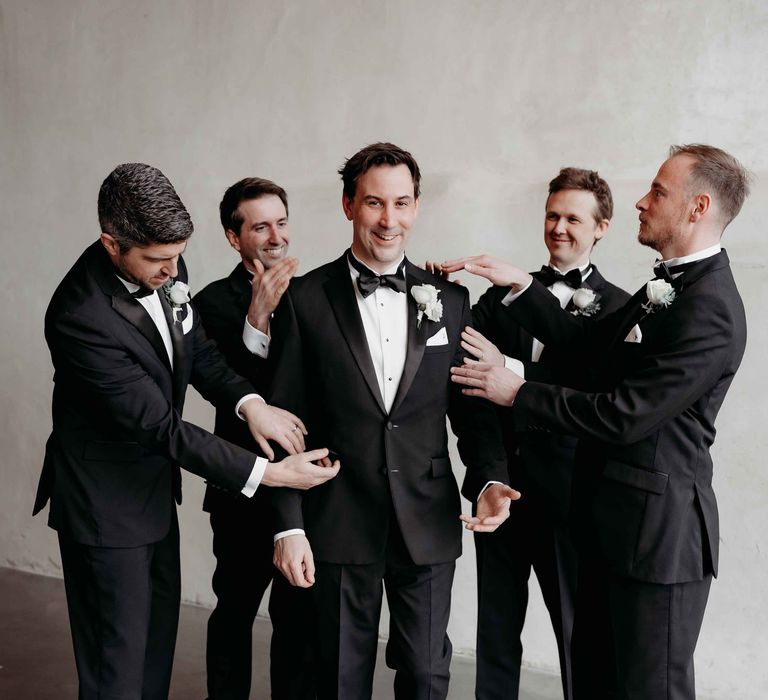 Groomsmen in black tuxedos fix up the groom in a black tuxedo and white rose boutonnière