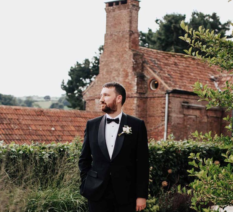 Groom walking around Brickhouse Vineyard Devon wedding venue wearing classic black grooms three piece tuxedo with white rose boutonniere and black bowtie 