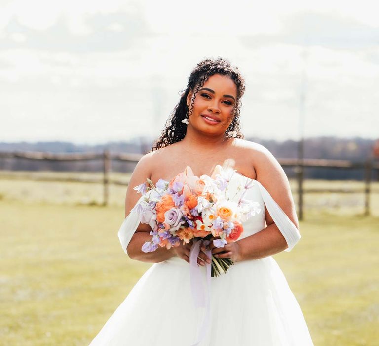 Bride in off the shoulder wedding dress and silver bridal crown holding orange, lilac and peach mixed flower bridal bouquet at Rackeys Barn