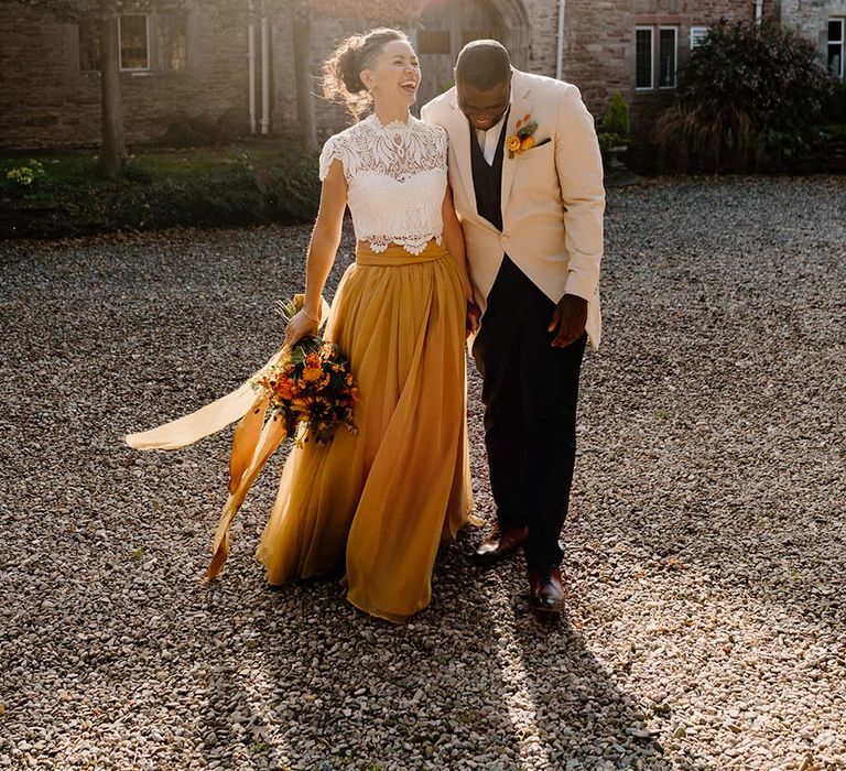 Groom in cream morning suit with forest green grooms waistcoat and trousers, forest green pocket square and orange boutonniere walking with bride in white mesh lace bridal crop top and yellow wedding skirt holding dried autumnal wedding bouquet with sunflowers, garden roses, pampas grass, eucalyptus, yellow tall kangaroo paws, orange osmanthus flowers, foliage and dried flowers at Broadfield Court